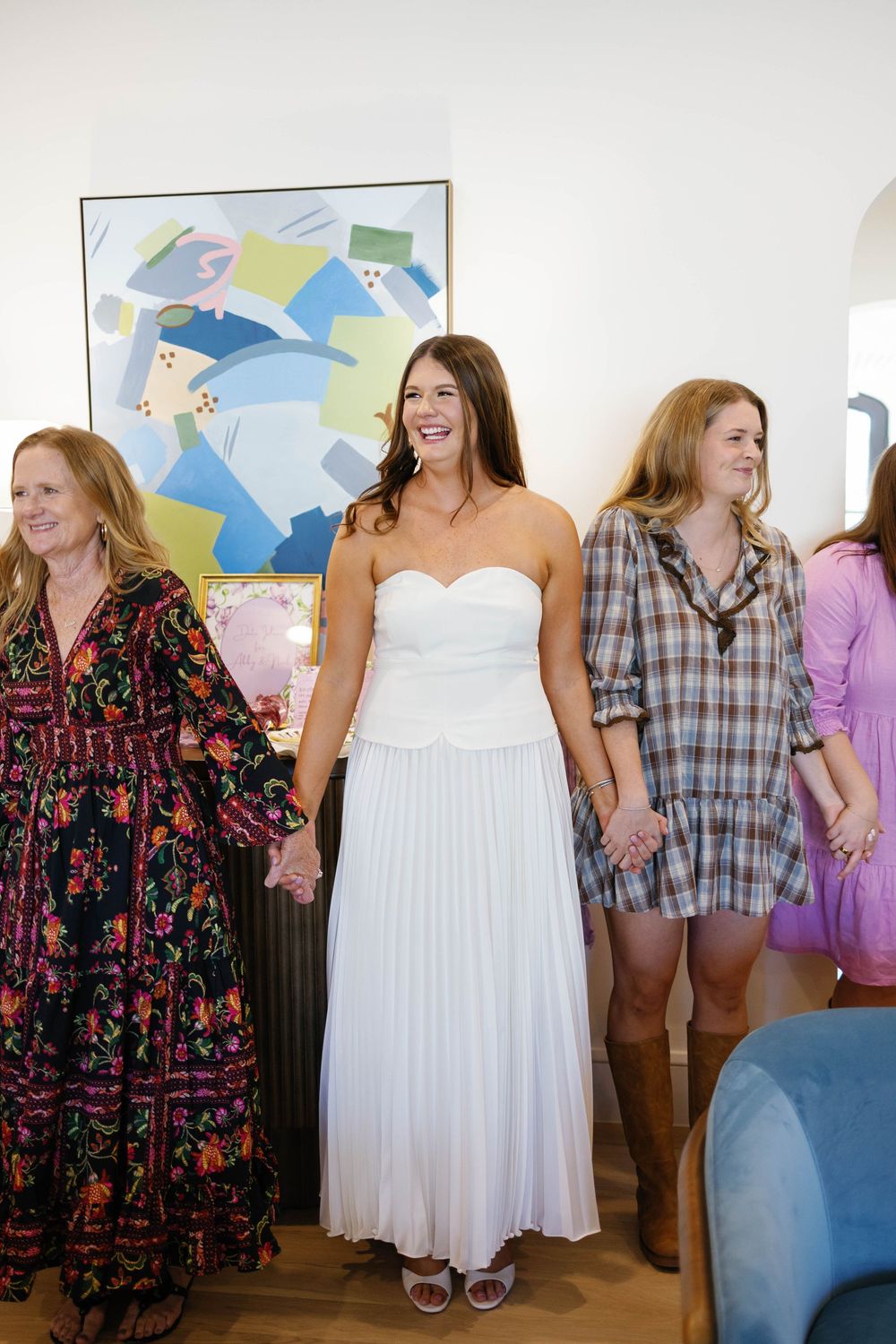 Bride in white strapless pleated gown holds hands with guests at bridal shower celebration indoors.