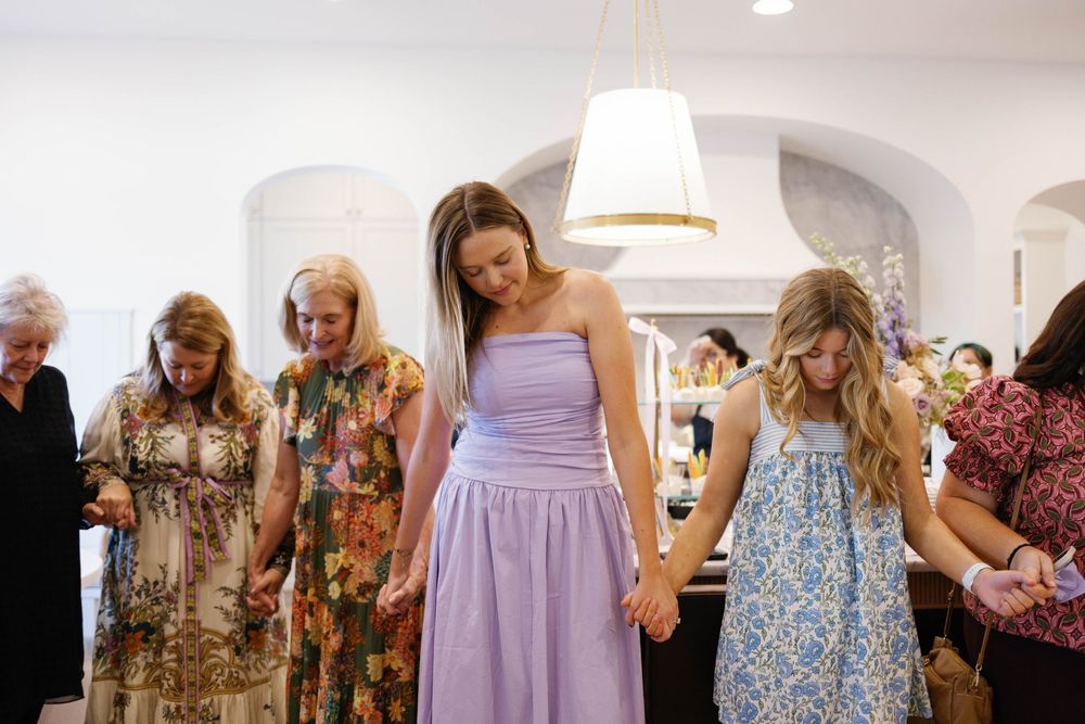 Women holding hands in prayer circle at a bridal shower event wearing colorful dresses.