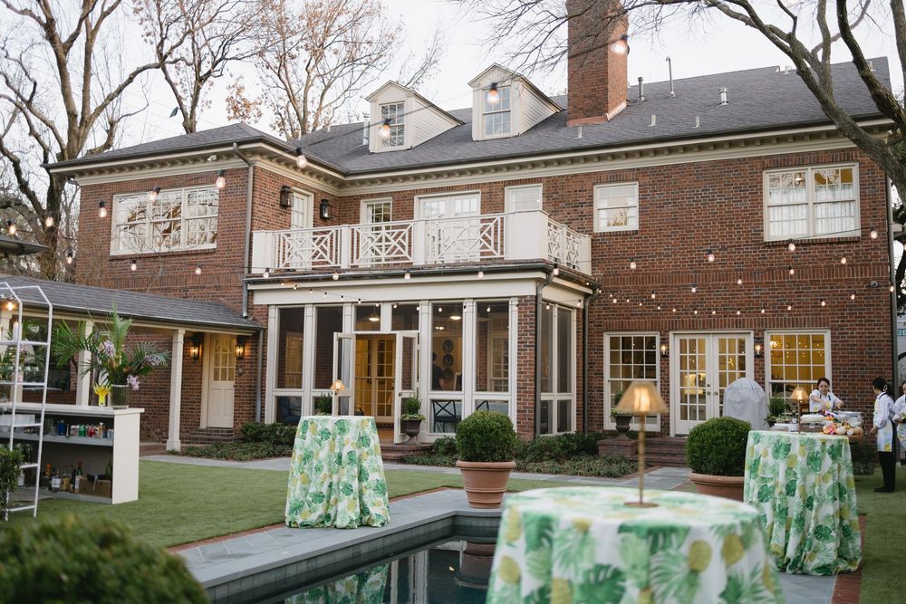 Elegant brick mansion with green and white floral tablecloths on cocktail tables in a manicured garden at dusk.