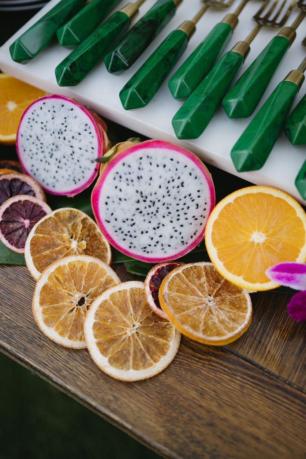 Sliced dragon fruit, dried orange rounds, and blood oranges displayed on a wooden table with green-handled cutlery.