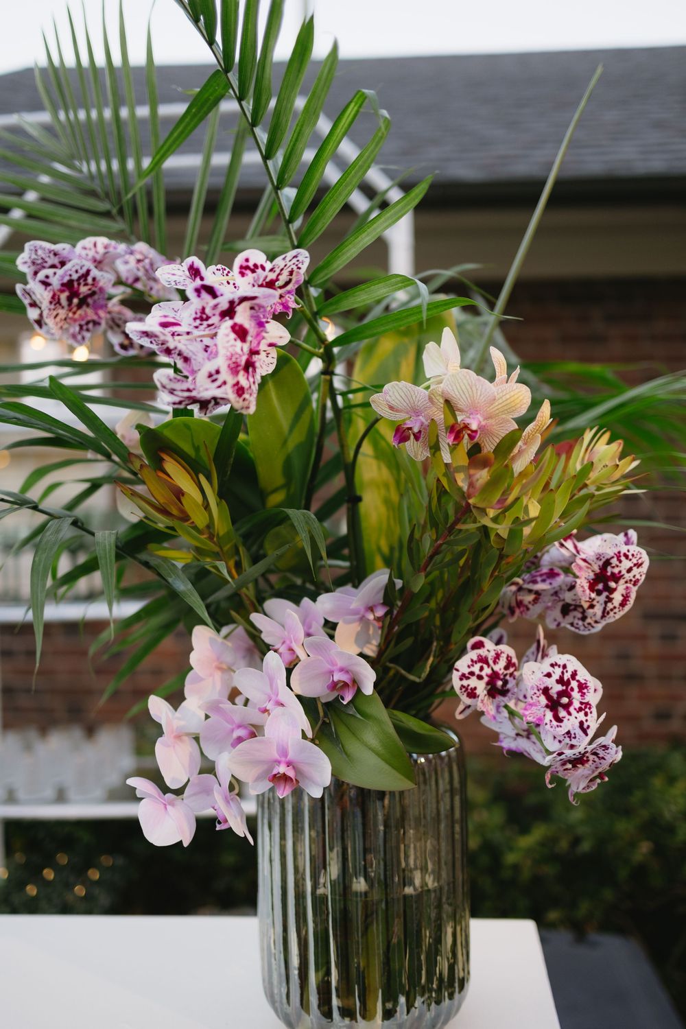Tropical floral arrangement featuring pink orchids, palm leaves, and protea in a ribbed glass vase outdoors.