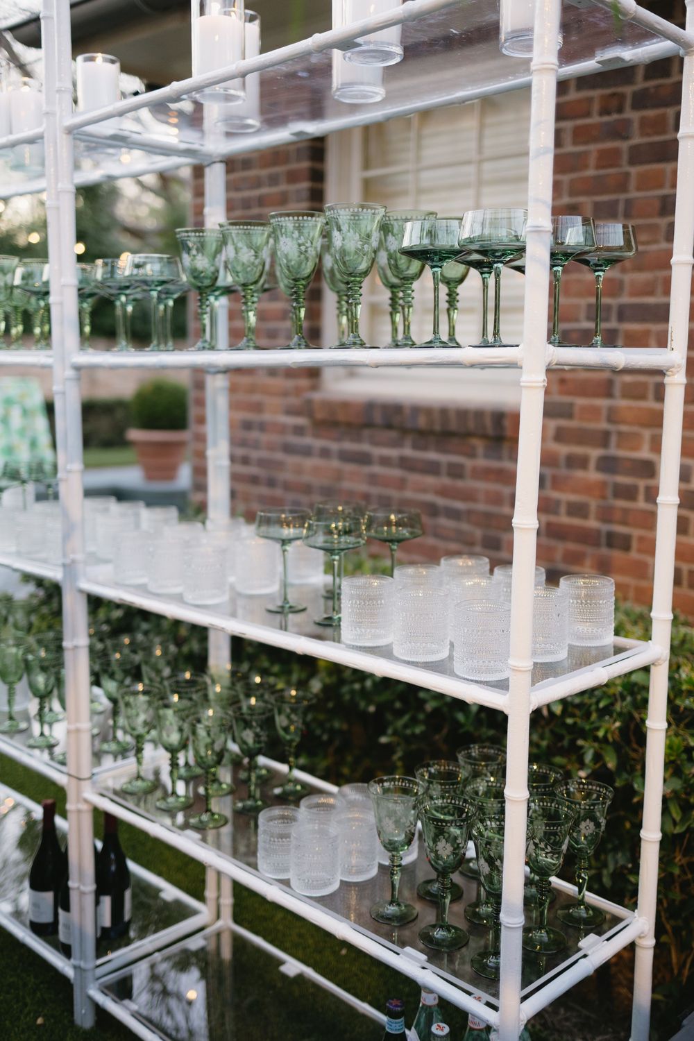 White shelving unit displaying green stemmed wine glasses and frosted tumblers against a brick wall backdrop.