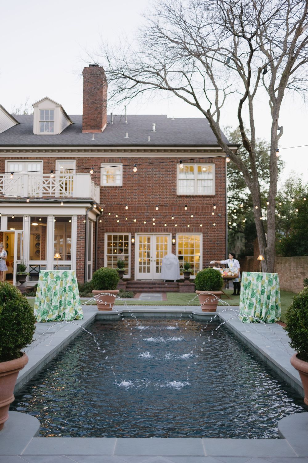 Classic brick estate with a rectangular reflecting pool, string lights, and tropical print cocktail tables at twilight.