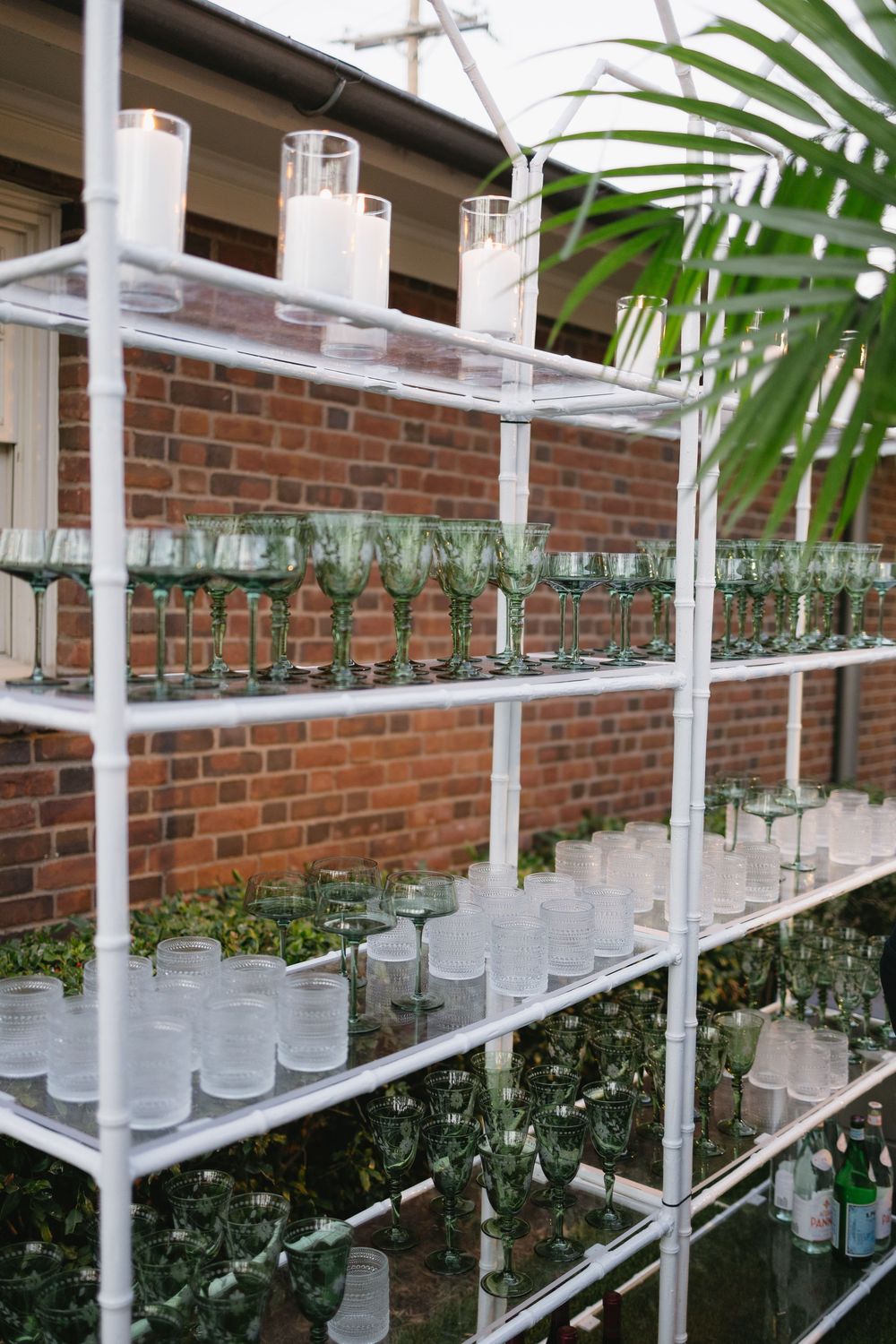 White metal shelving unit with green wine glasses, frosted tumblers, and pillar candles against a brick wall.