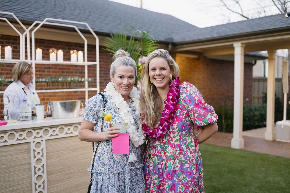 Two smiling women in colorful floral dresses wearing leis at an outdoor tropical-themed garden party.