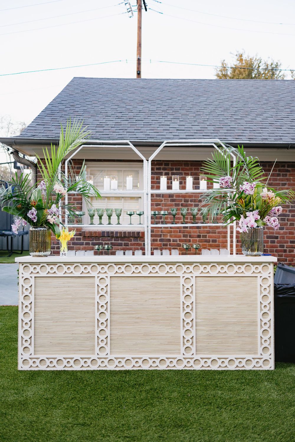Decorative outdoor bar setup with tropical floral arrangements and green glassware on manicured lawn.