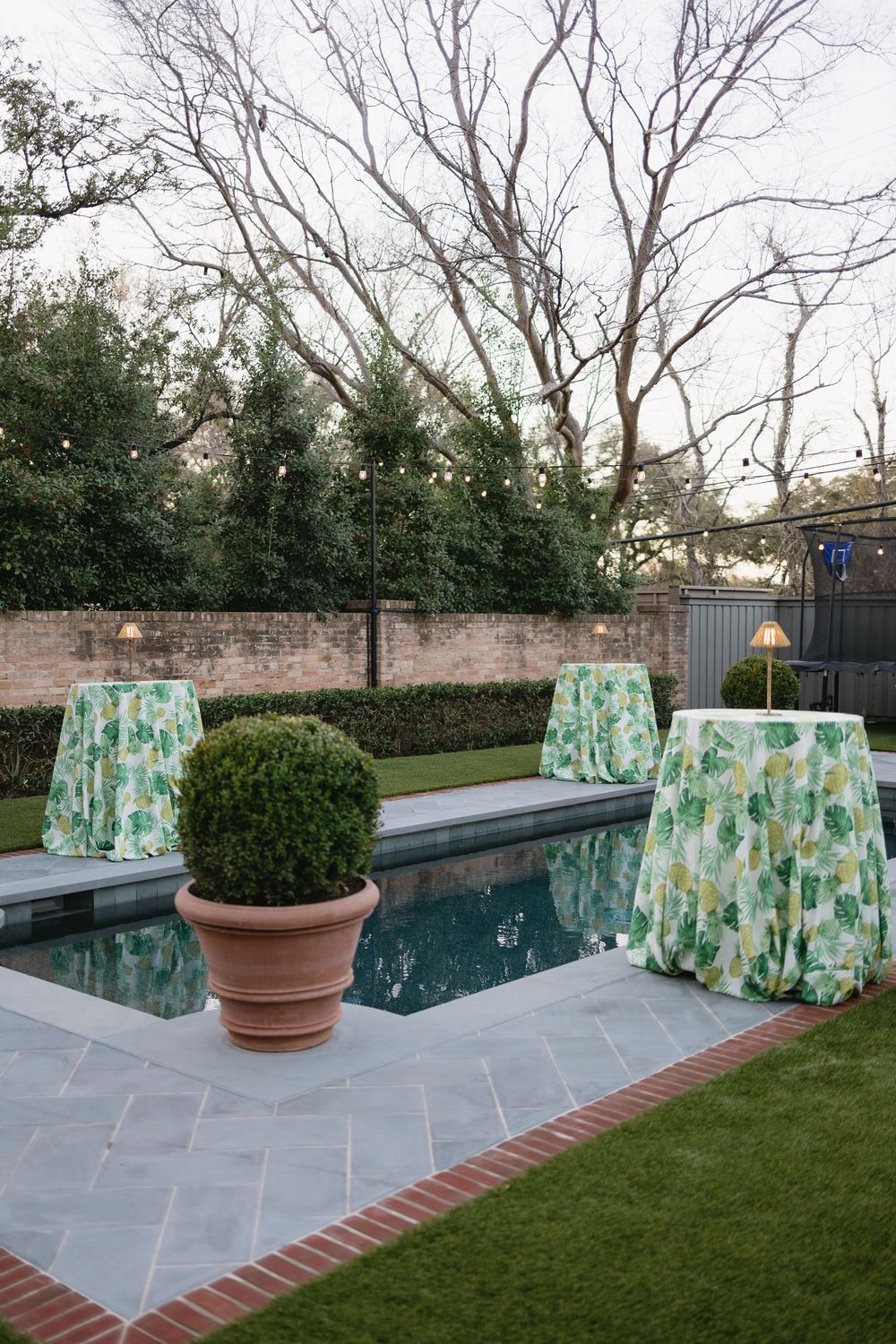 Poolside cocktail hour setup with green floral tablecloths, boxwood topiary, and string lights at dusk.