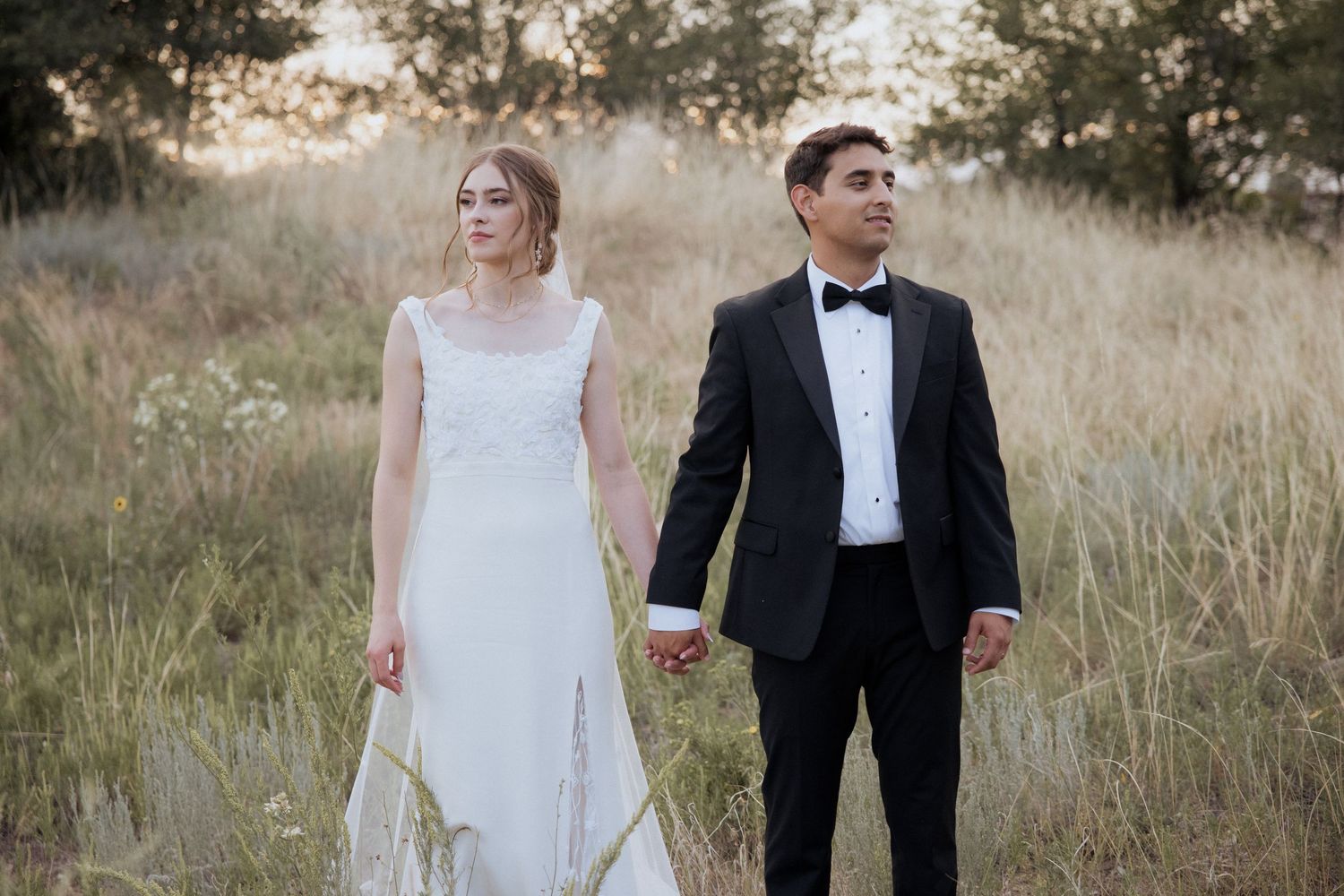 A couple in wedding attire stands together on a garden path surrounded by trees and greenery.