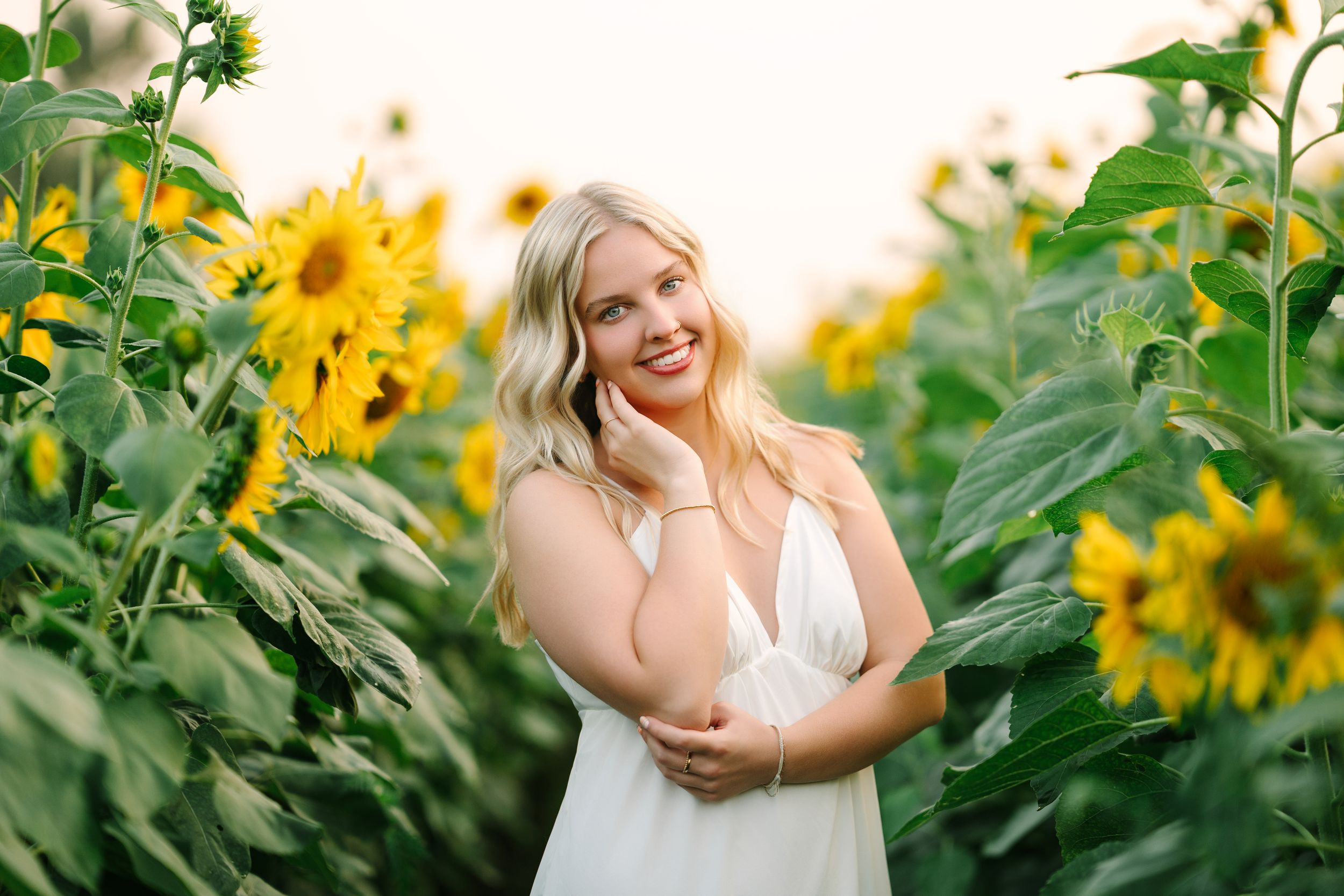 Smiling blonde woman in white dress standing among tall sunflowers in a sunny field.