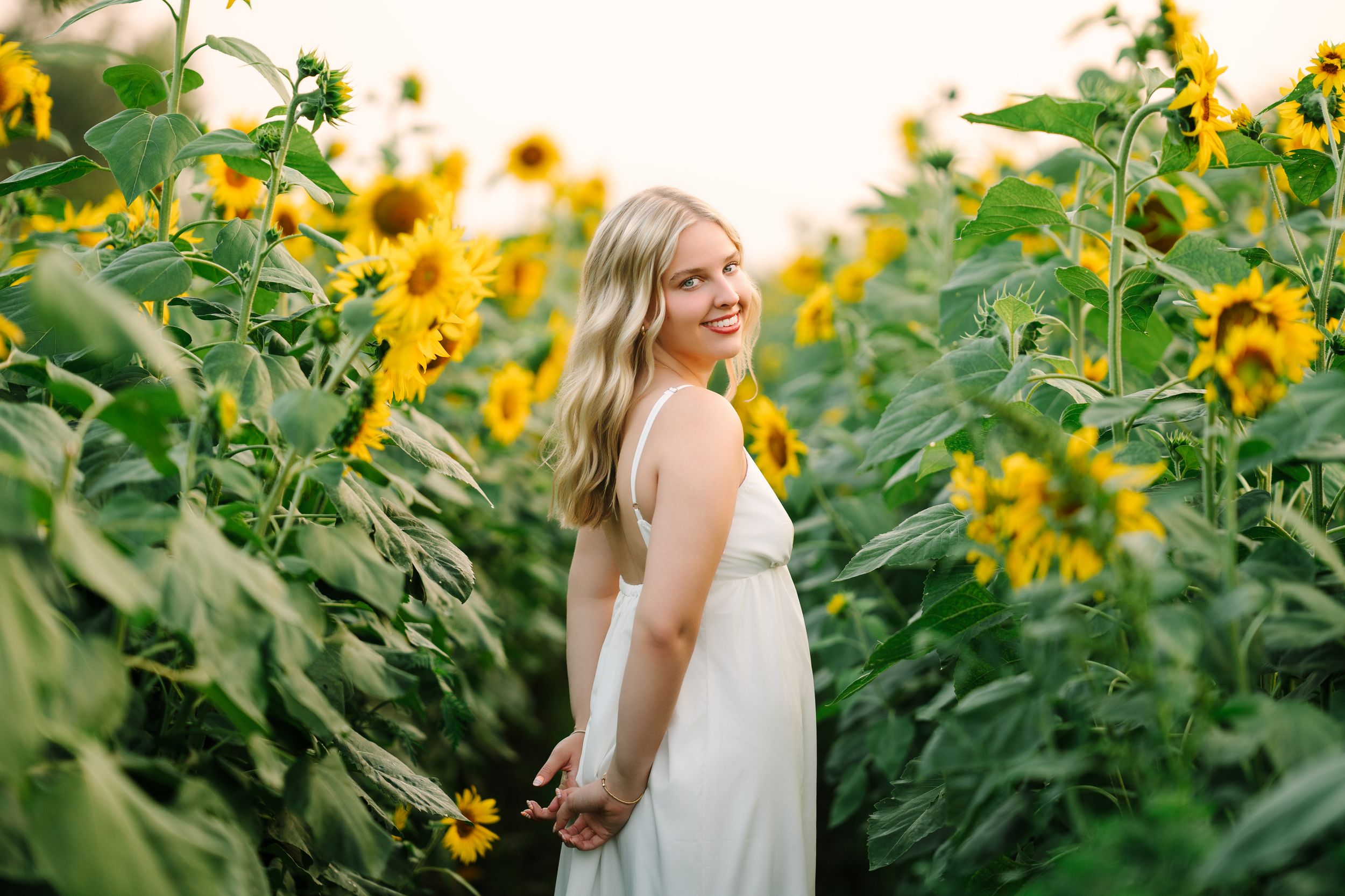 Young blonde woman in white dress smiling among tall sunflowers in a sunny field.