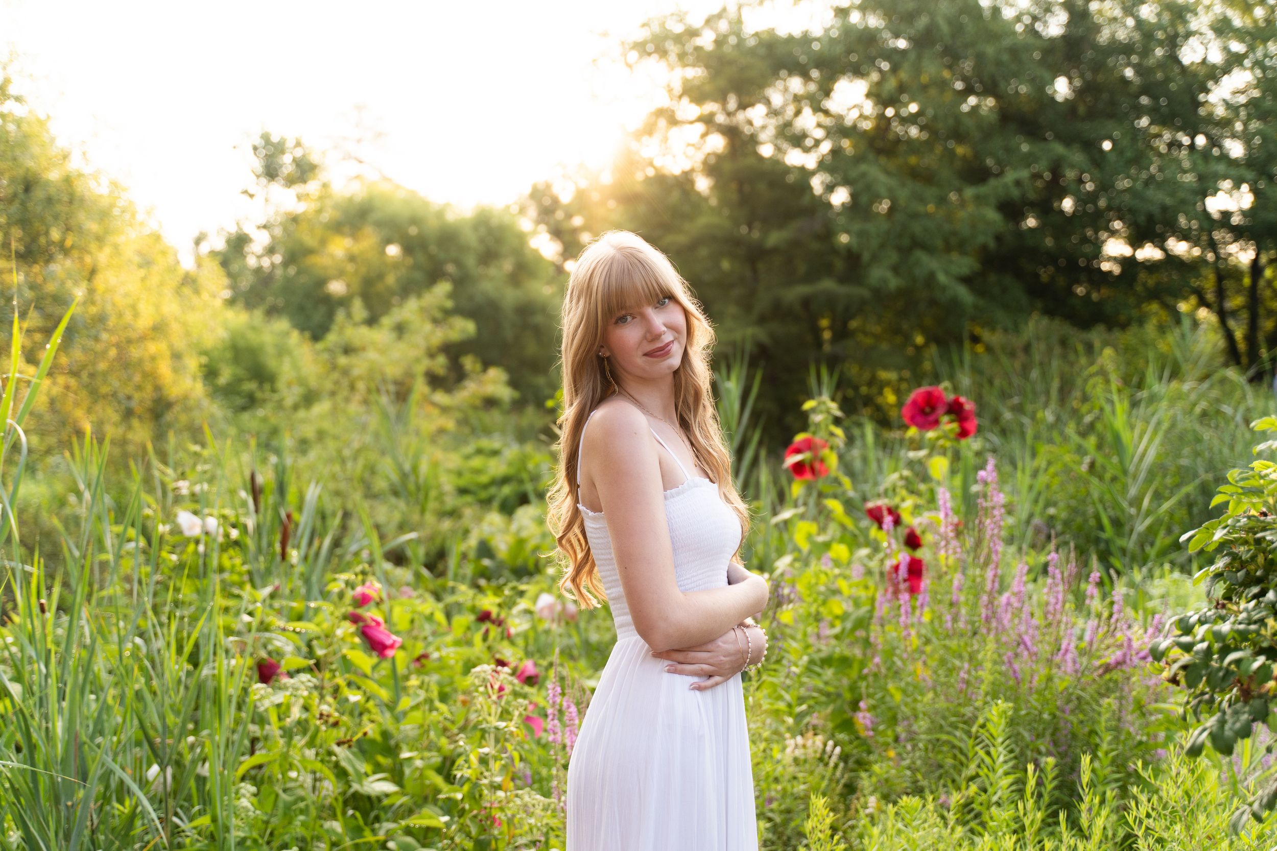 A Perrysburg High School senior in a white dress stands among vibrant wildflowers during golden hour in a summer garden.