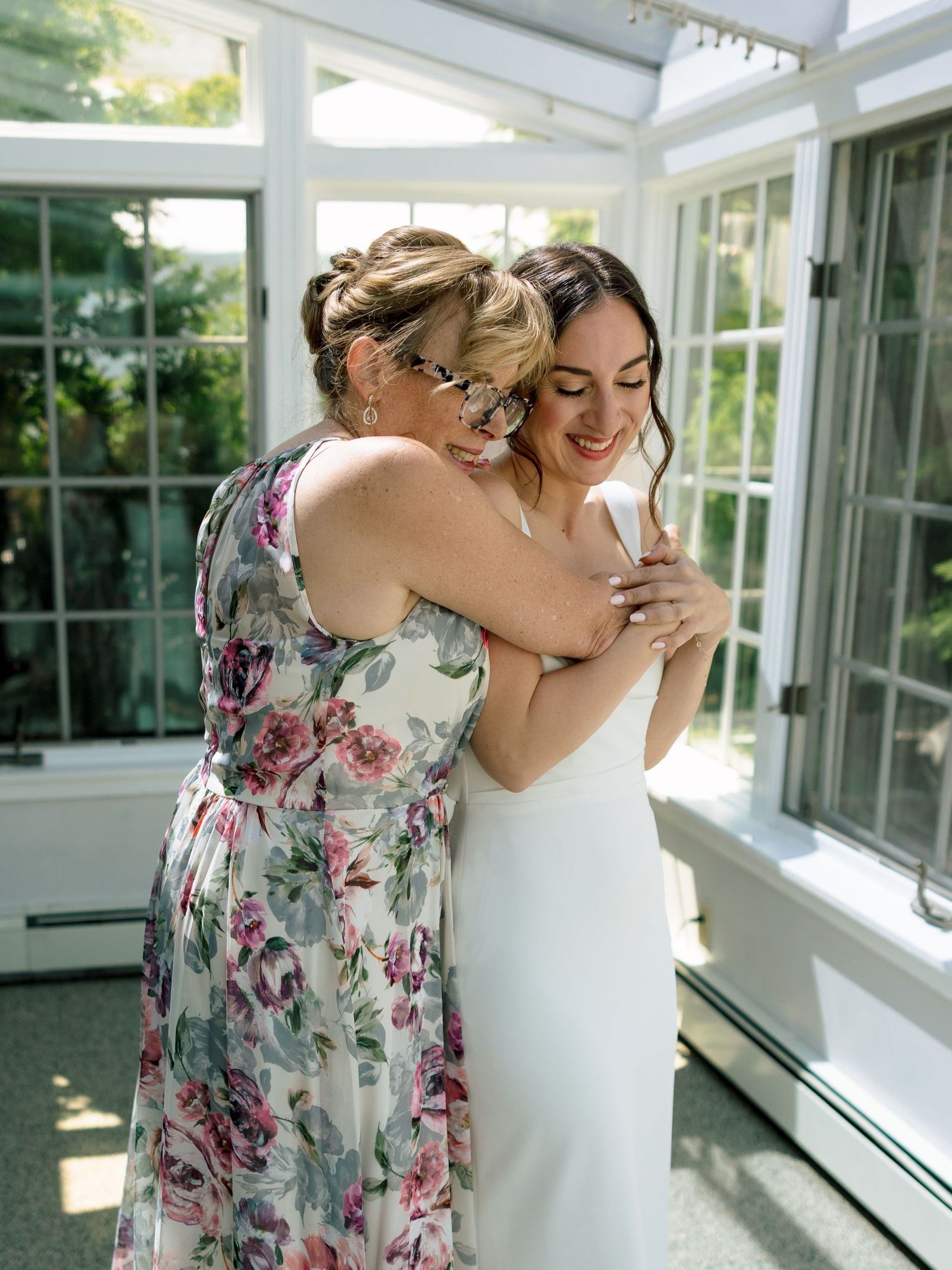 Mother and bride share an embrace in a sunlit conservatory, one wearing a floral dress and one in a white satin bridal gown.