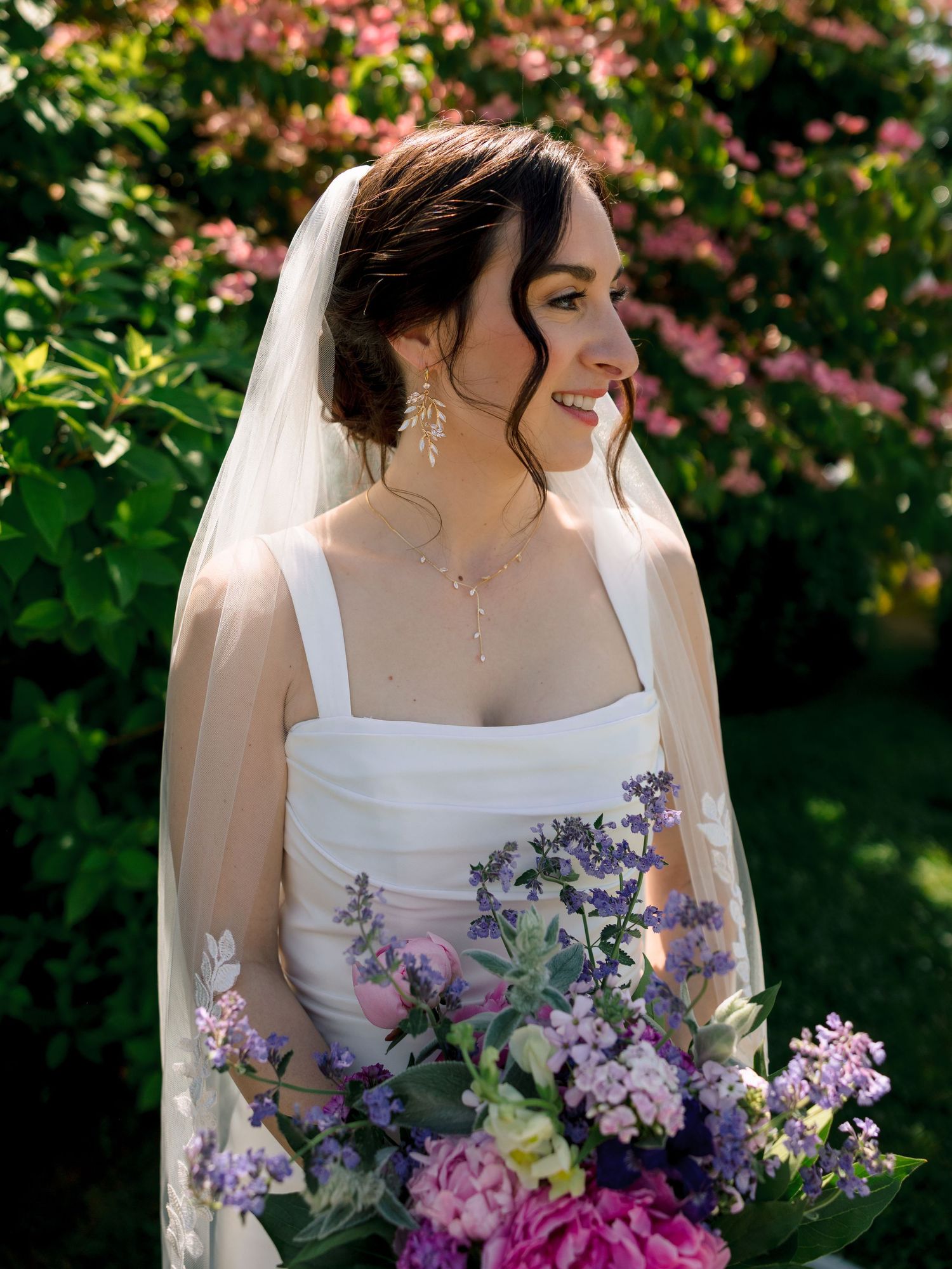 A bride in a white dress holding a vibrant purple and pink bouquet against a garden backdrop with pink blossoms.