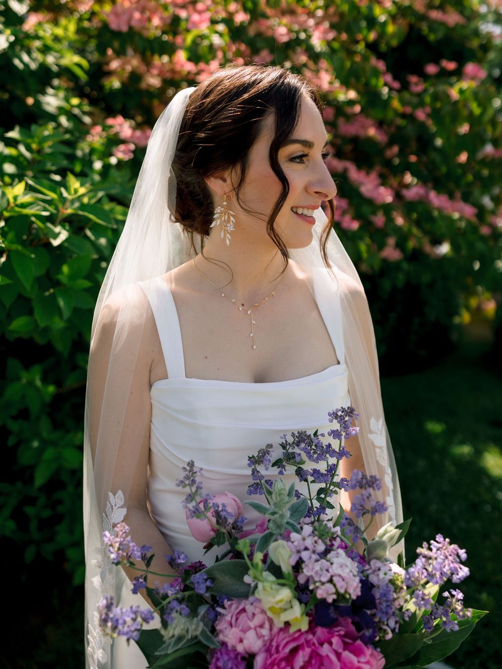 A bride in a white dress holding a vibrant purple and pink bouquet against a garden backdrop with pink blossoms.