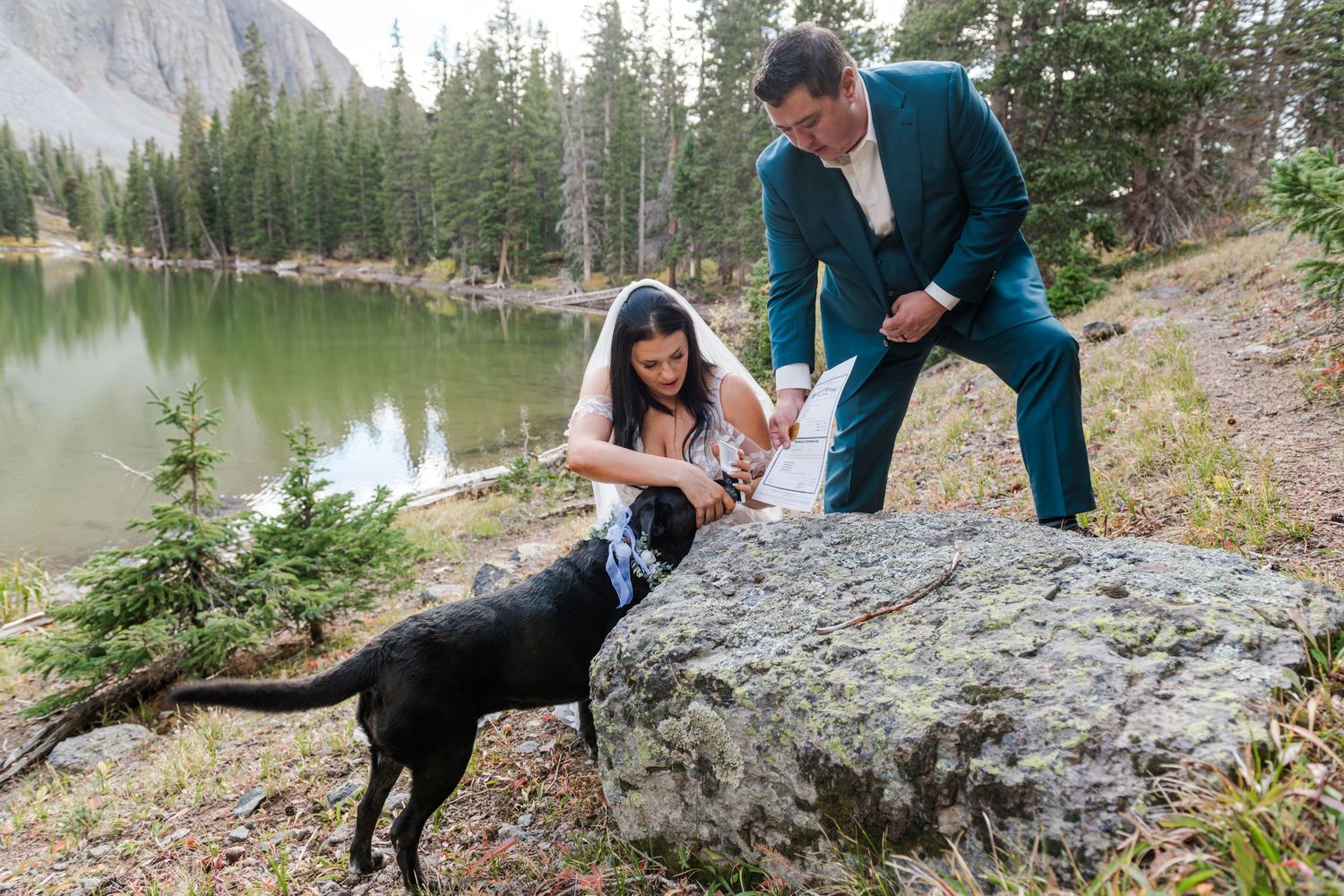 A bride and groom help their dog to sign the marriage license with his nose near an alpine lake elopement ceremony.