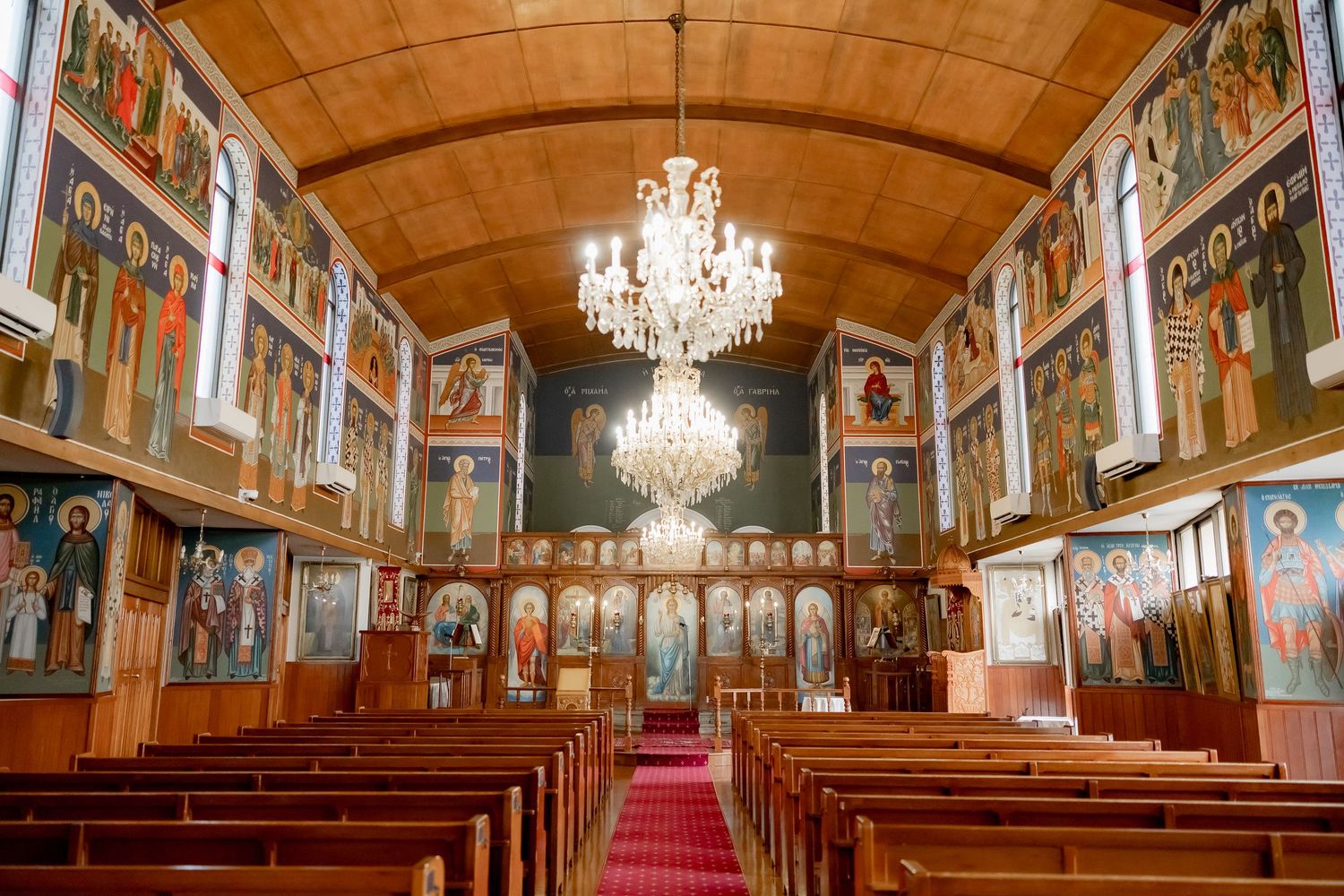 Panoramic view of an ornate church interior with crystal chandeliers, wooden pews, and religious artwork along the walls.