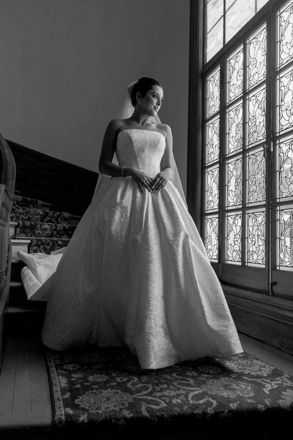 Dramatic black and white portrait of a bride in a ballgown next to a window.