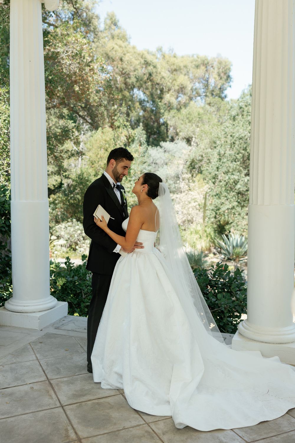 Romantic wedding couple embracing between white columns with lush garden backdrop.