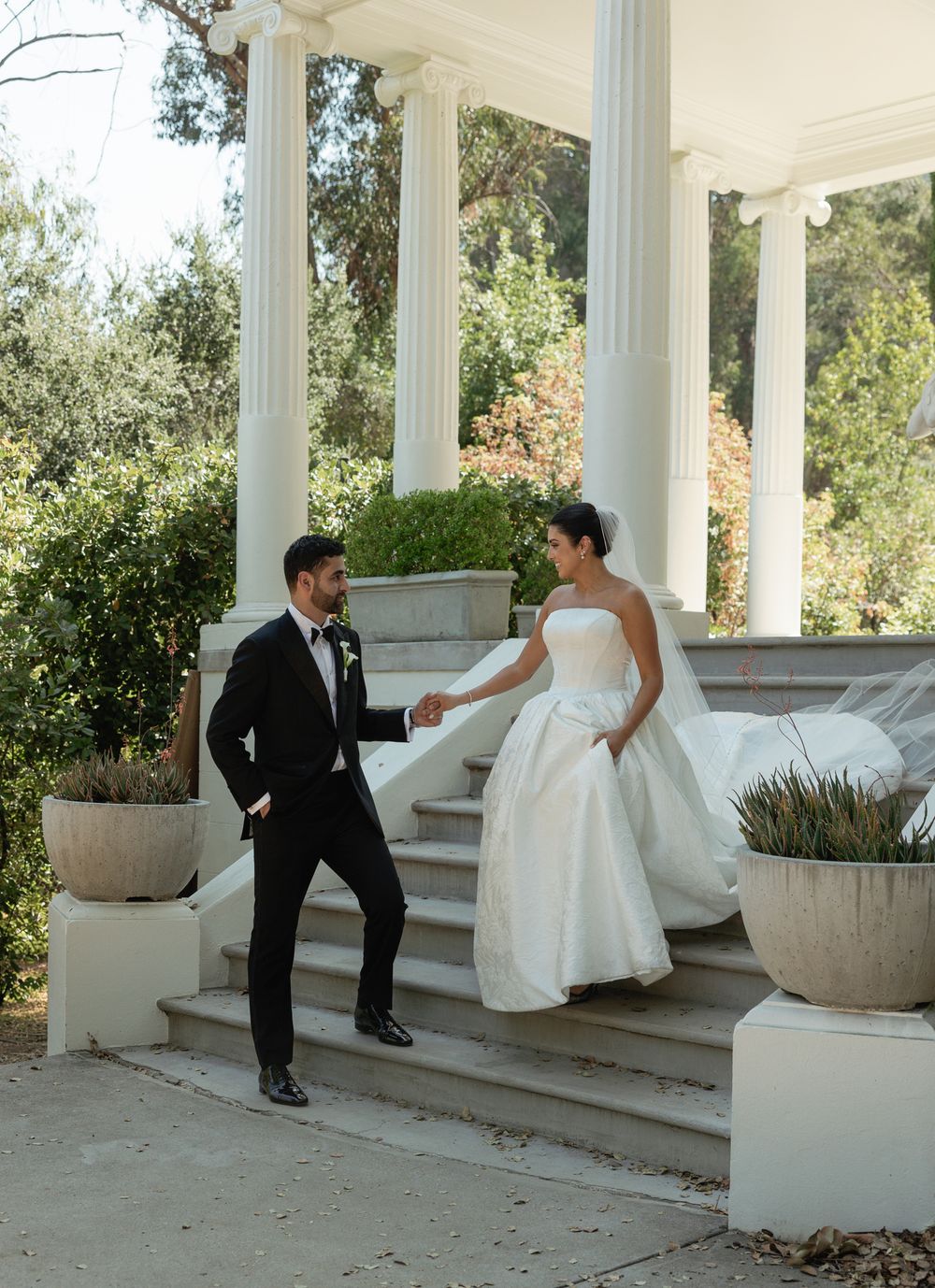 Playful wedding couple interaction on white columned steps with garden views.
