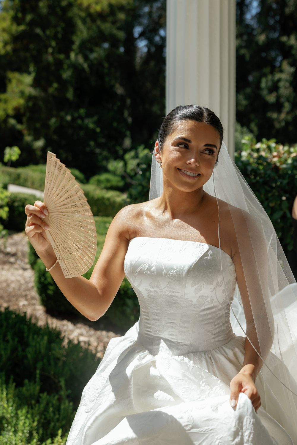 A bride in a white wedding dress poses with a decorative fan in an outdoor garden setting.