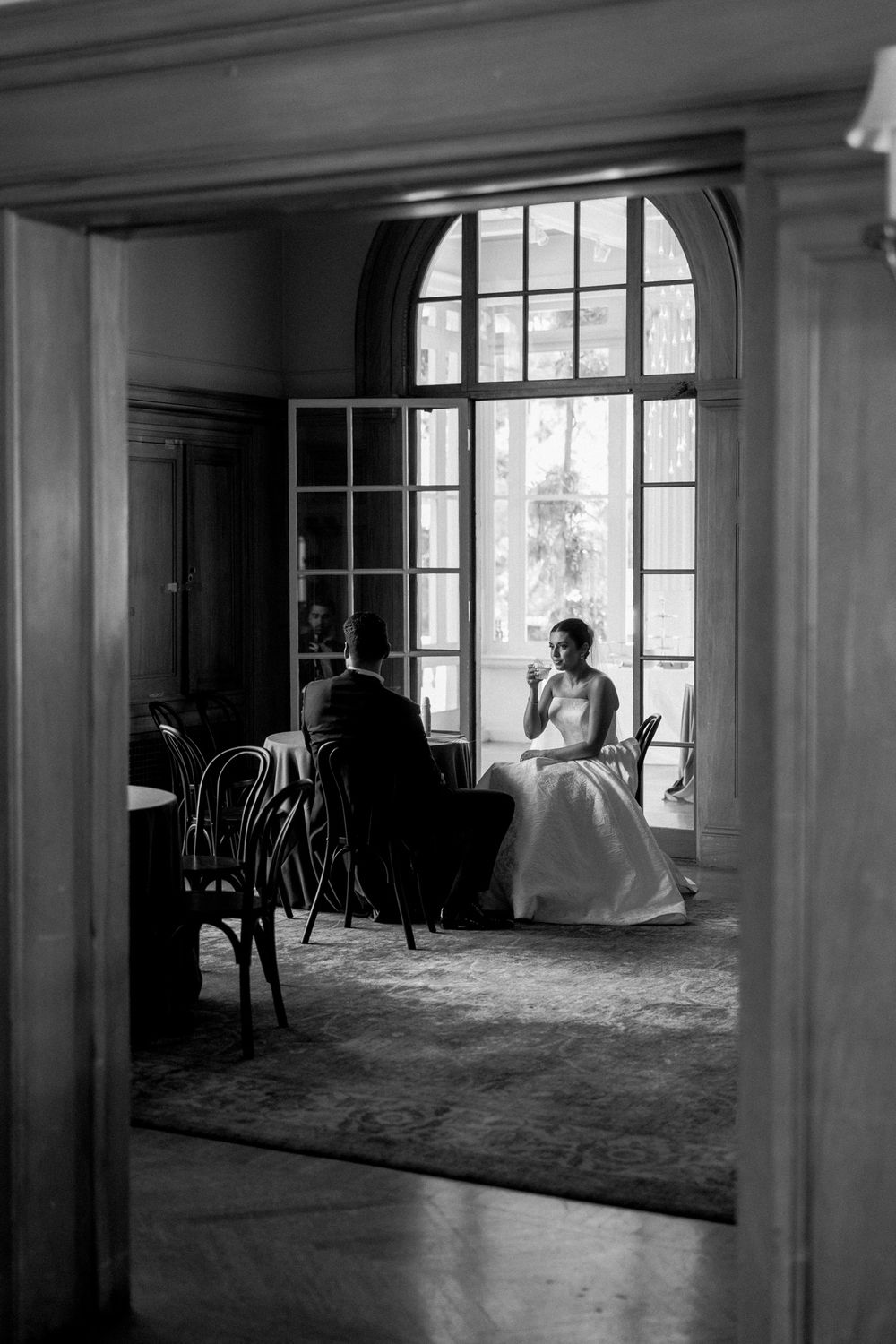 Black and white photo of an elegant room with large windows and scattered chairs.
