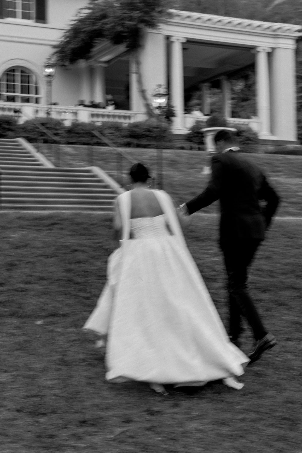 Couple dances and spins across lawn with historic building backdrop at wedding celebration.