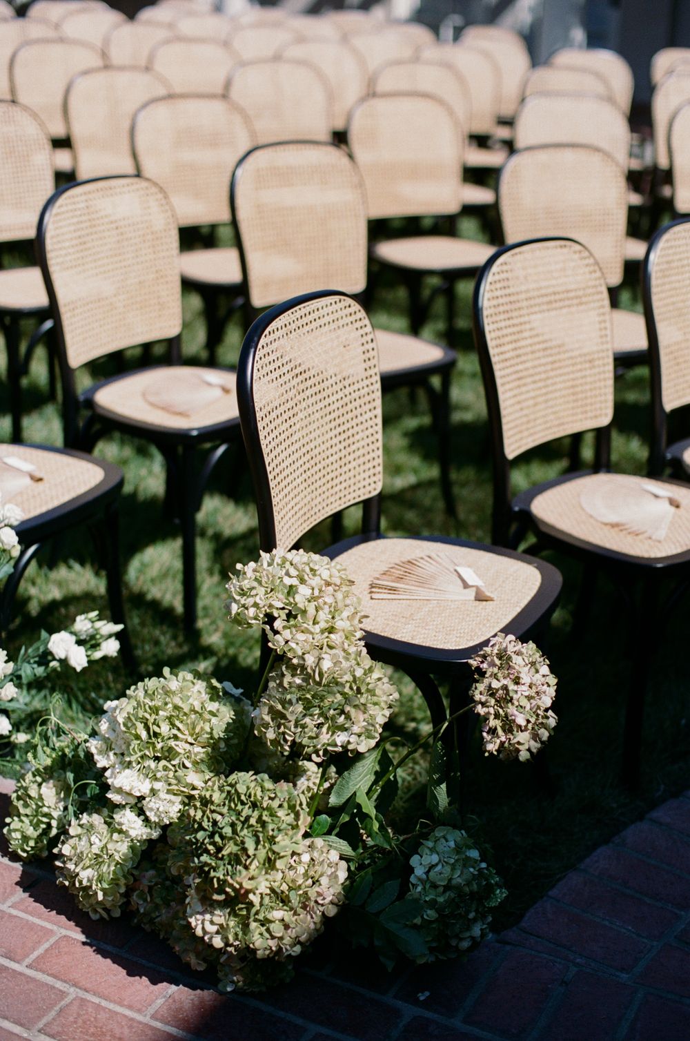 Empty cane chairs arranged for outdoor wedding ceremony with hydrangea floral decorations.
