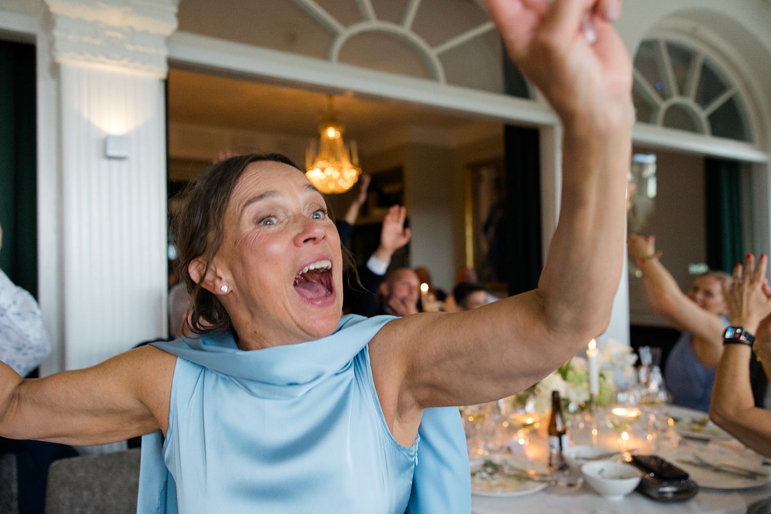 A person in a light blue shirt celebrates joyfully with raised arms at a dining table during a gathering.