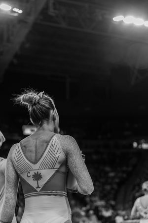 Black and white photo of a female gymnast in a decorative leotard, viewed from behind in a competition arena.