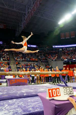 Clemson gymnast performs a leap on the balance beam during a packed arena competition, with scores 100 and 985 displayed.
