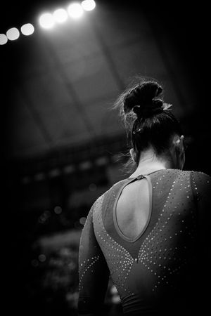Black and white photo of a female gymnast in a rhinestone leotard with hair in a bun, facing a bright arena.