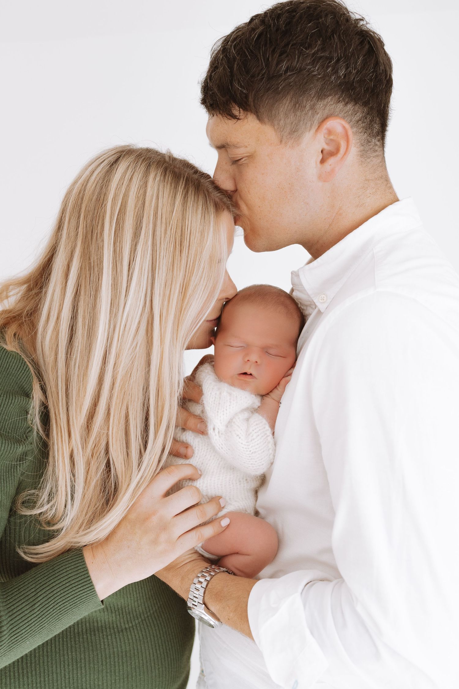 Parents in green and white tops share tender moment while holding their sleeping newborn baby wrapped in white.