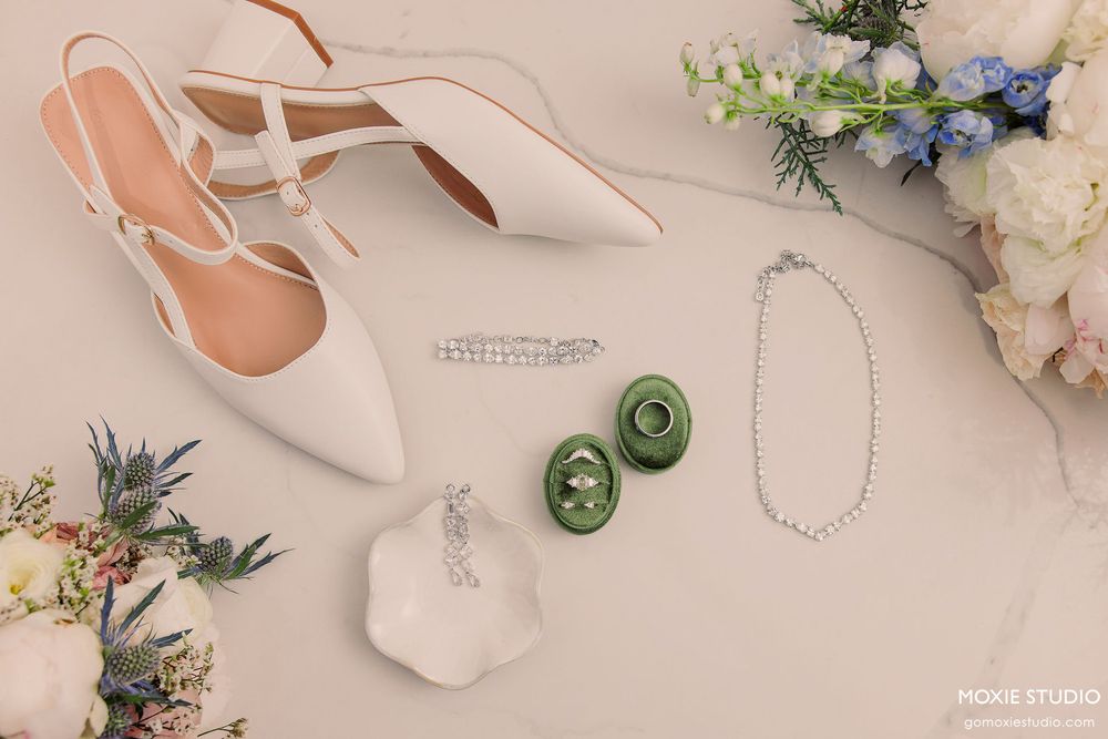 Elegant white bridal accessories including slingback heels, jewelry, and floral arrangements on beige background.