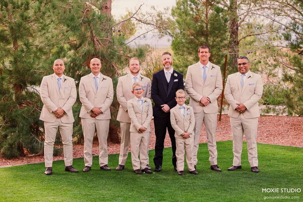 Wedding party in khaki suits sits casually on a wooden bench in an outdoor garden setting at sunset.