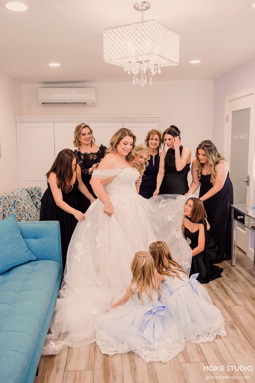A group in black dresses and white wedding gowns gather in a modern room with light wood floors and a blue couch.