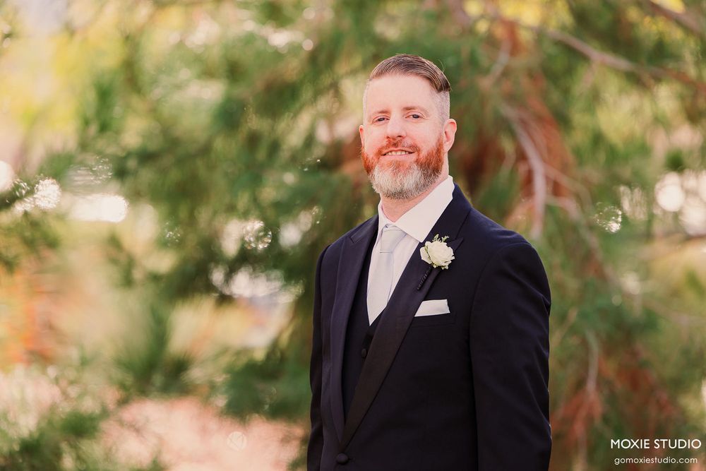 A groom in a black suit and tie smiles outdoors with autumn foliage creating a warm, natural backdrop.