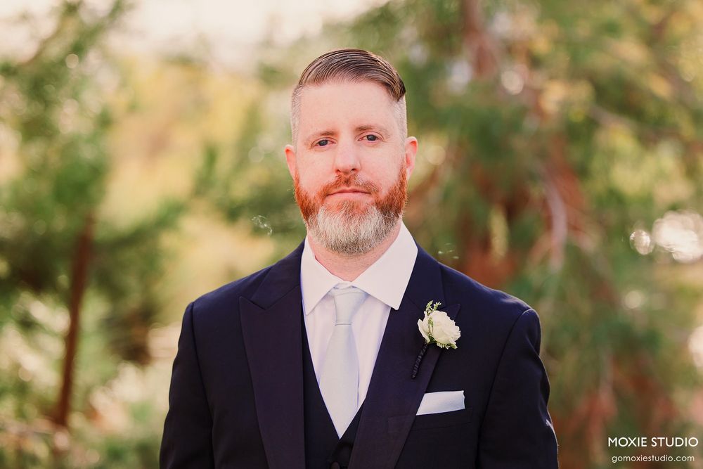 Groomsmen in black suits and white ties stand together in a garden setting during a formal wedding event.