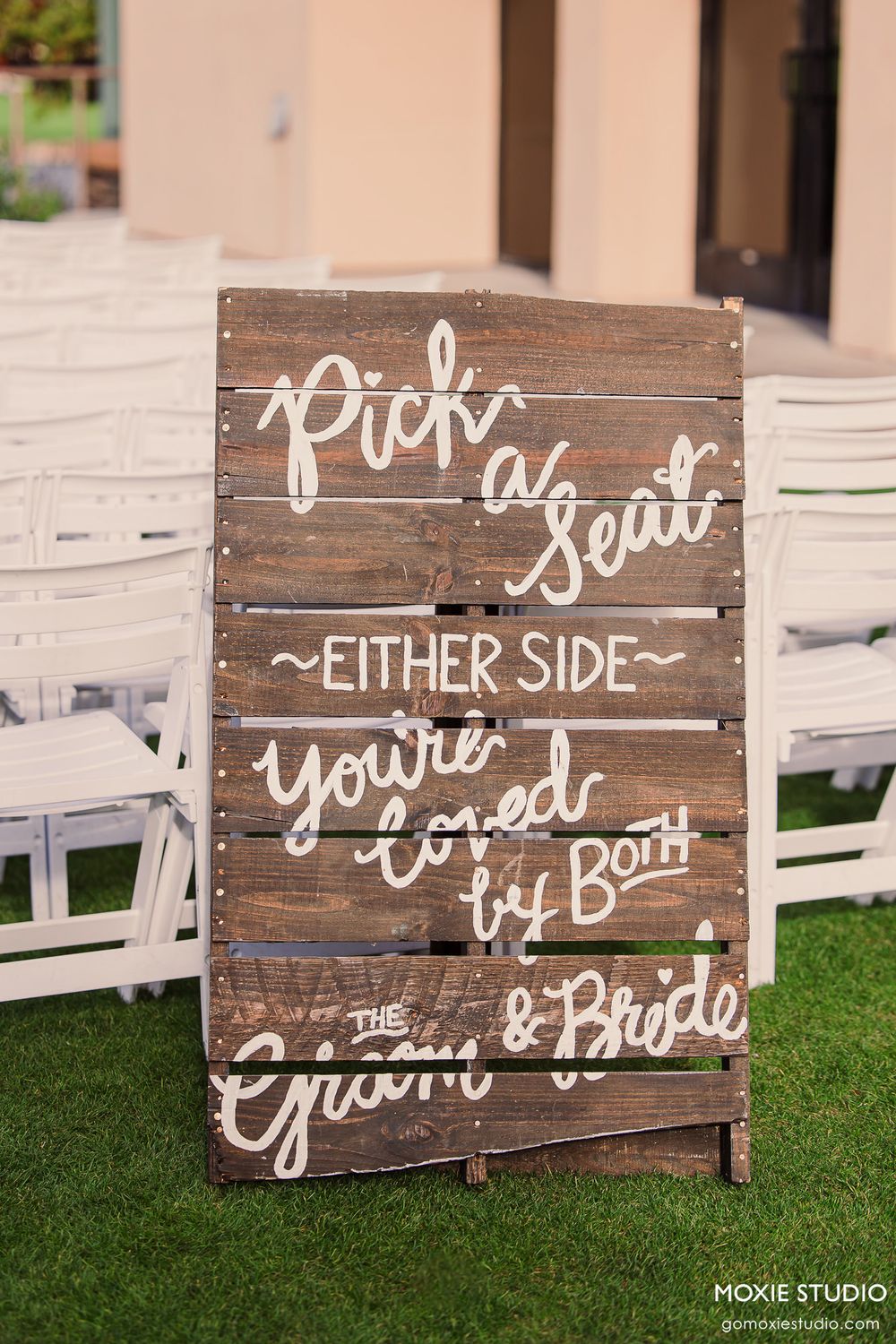 Rustic wooden wedding signs with white handwritten text displayed on grass at an outdoor ceremony setup.