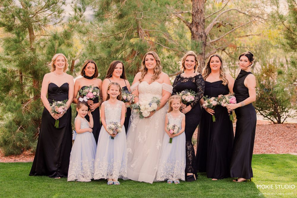 Wedding party in elegant black gowns and white dresses pose together on grass with pine trees in background.