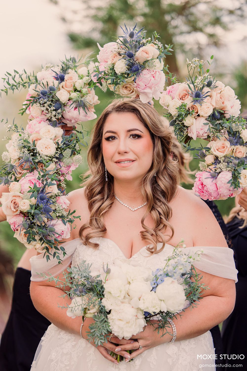 Bride in off-shoulder wedding dress holds elegant bouquet of peach and white flowers with blue accents and greenery.