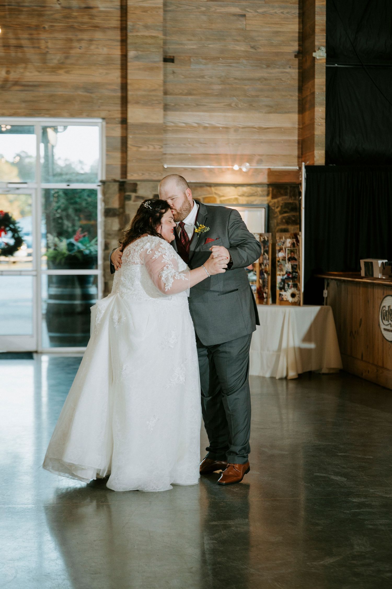 A couple shares an intimate dance moment in a rustic wooden-walled reception venue with natural lighting.