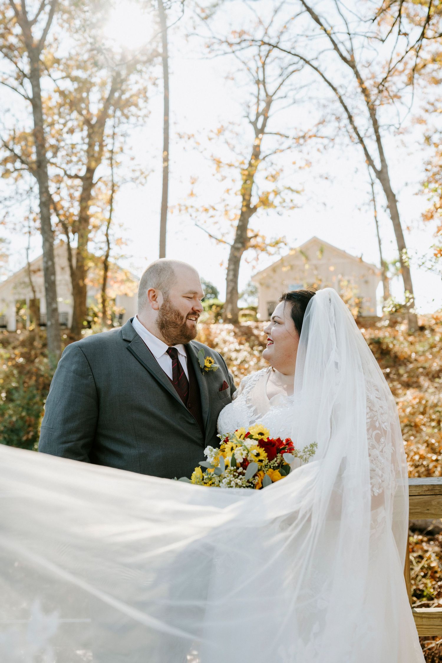 A couple poses together outdoors during autumn with sunlit trees and flowing white veil in the background.
