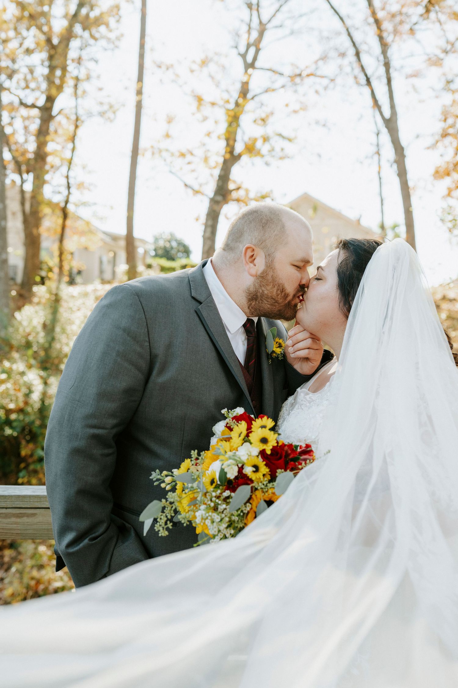 A romantic wedding couple shares intimate moments outdoors with flowing white veil and yellow bouquet in autumn setting.
