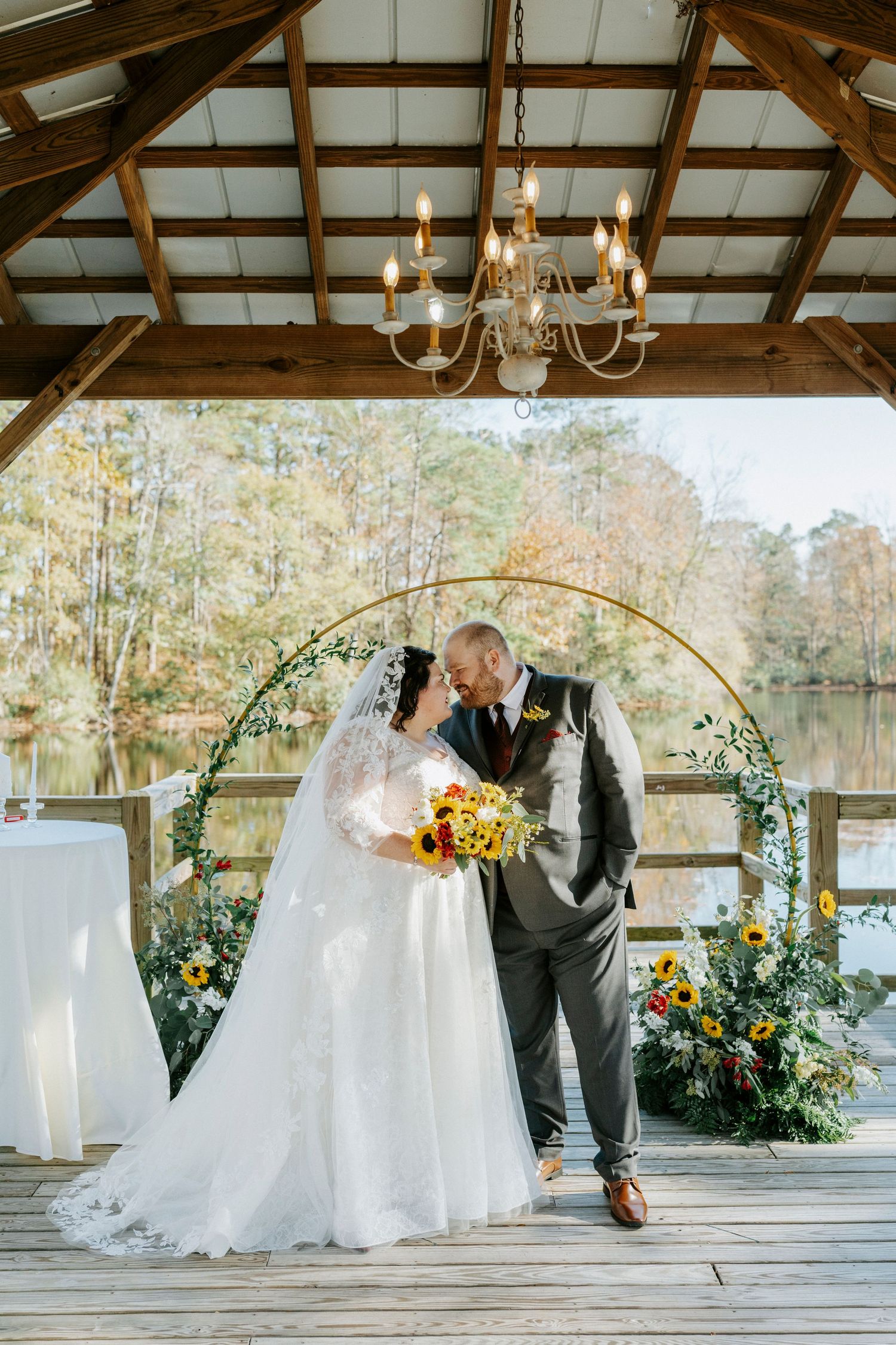 Romantic outdoor wedding ceremony in rustic pavilion with wooden beams and chandeliers overlooking lake in autumn.