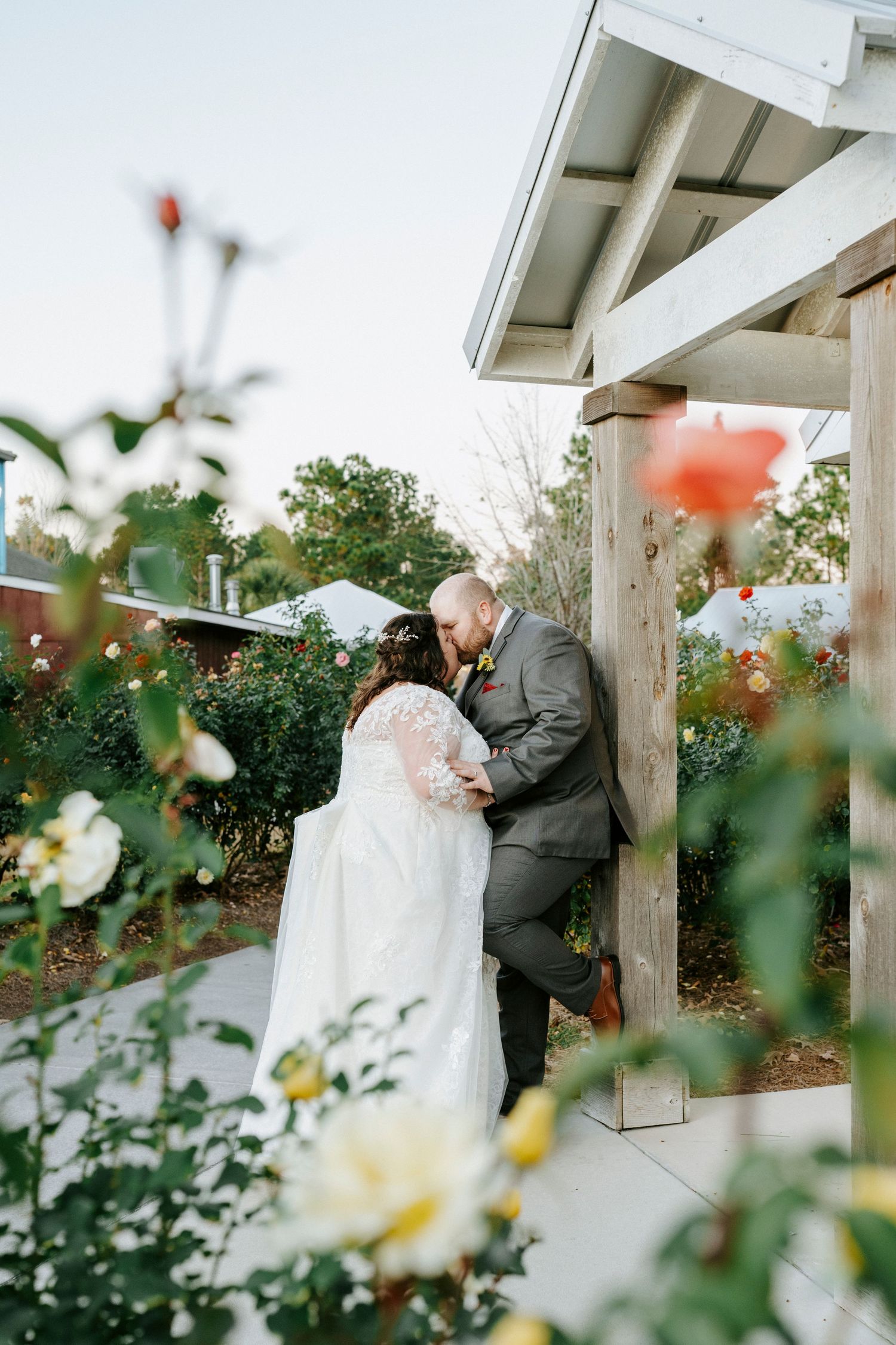A romantic wedding moment captured through blooming flowers in the foreground at an outdoor venue with white pillars.