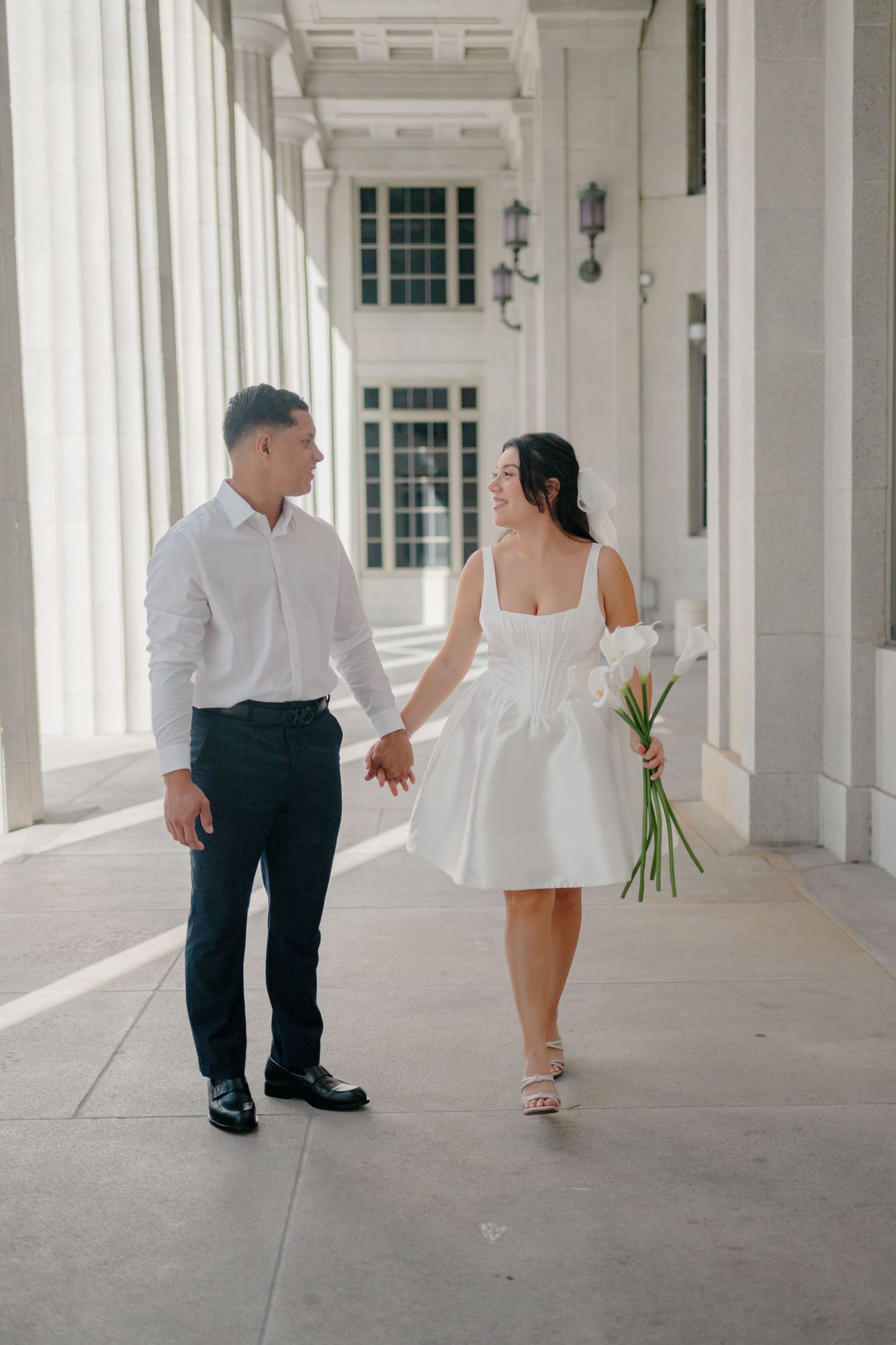A couple walks together down an elegant white columned hallway in formal attire.