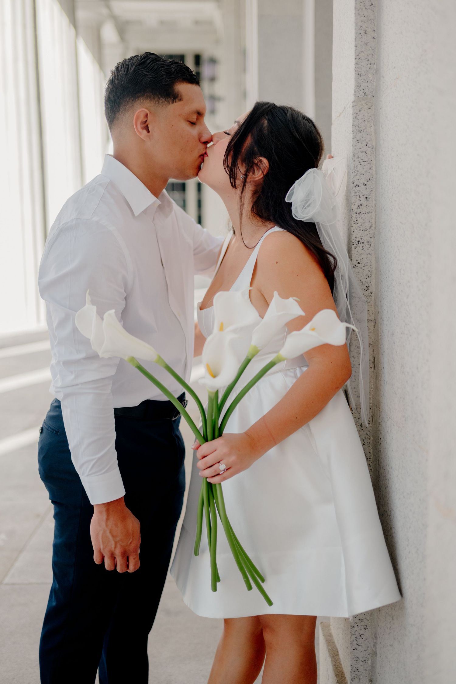 A romantic couple shares a tender kiss while holding white calla lilies in an elegant white hallway.