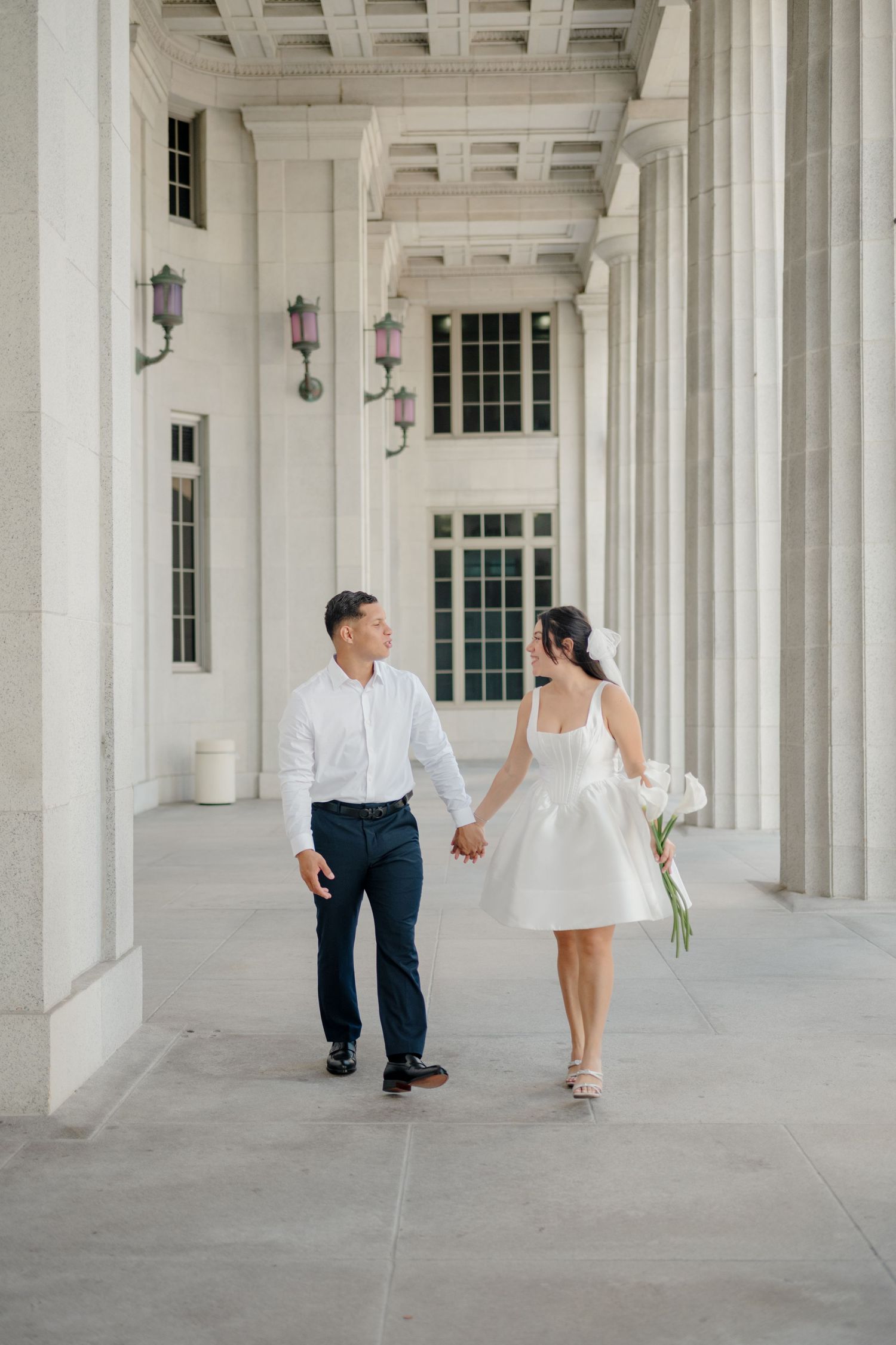 A couple walks hand in hand through an elegant white colonnade hallway with repeating architectural columns and arches.