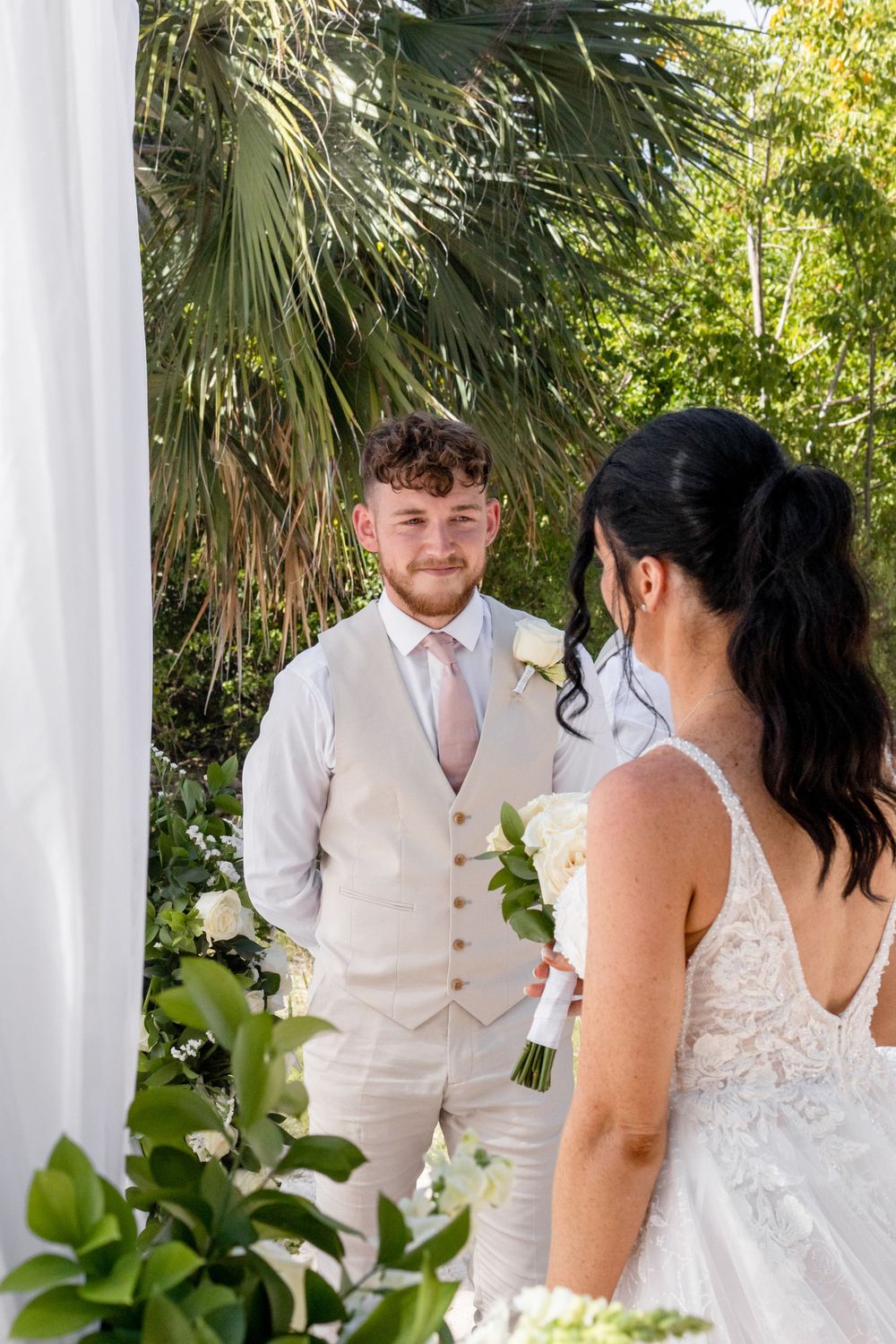 A couple shares a tender moment during their tropical wedding ceremony with palm trees in the background.