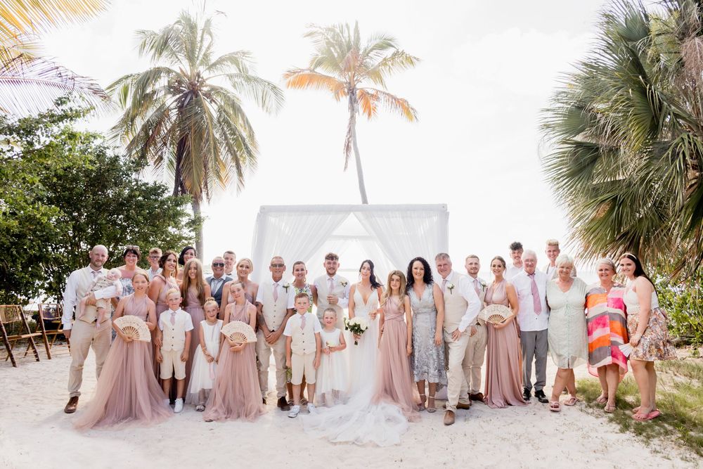 Wedding party in blush and neutral tones poses together on a tropical beach with palm trees swaying in the breeze.